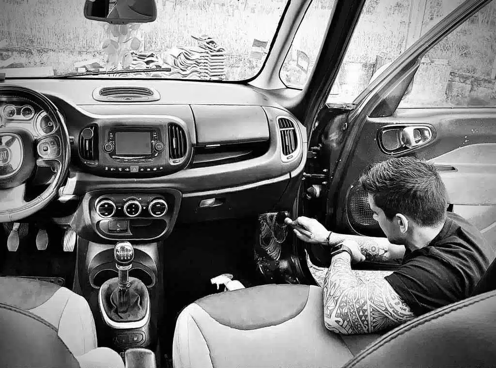 Nettoyage intérieur Fiat 500L : homme tatoué détaille la portière Homme tatoué nettoyant l'intérieur de portière d'une Fiat 500L, tableau de bord visible, vue depuis l'arrière.