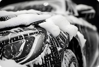 Lavage Voiture Mousseuse : Détail Phare Brillant Gros plan noir et blanc sur l'avant d'une voiture de luxe recouverte de mousse et de gouttes d'eau lors d'un lavage auto.