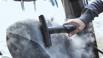 Nettoyage Vapeur Siège Voiture : Désinfection Efficace Nettoyage vapeur intense d'un siège de voiture en tissu gris avec une brosse, dégageant beaucoup de vapeur.